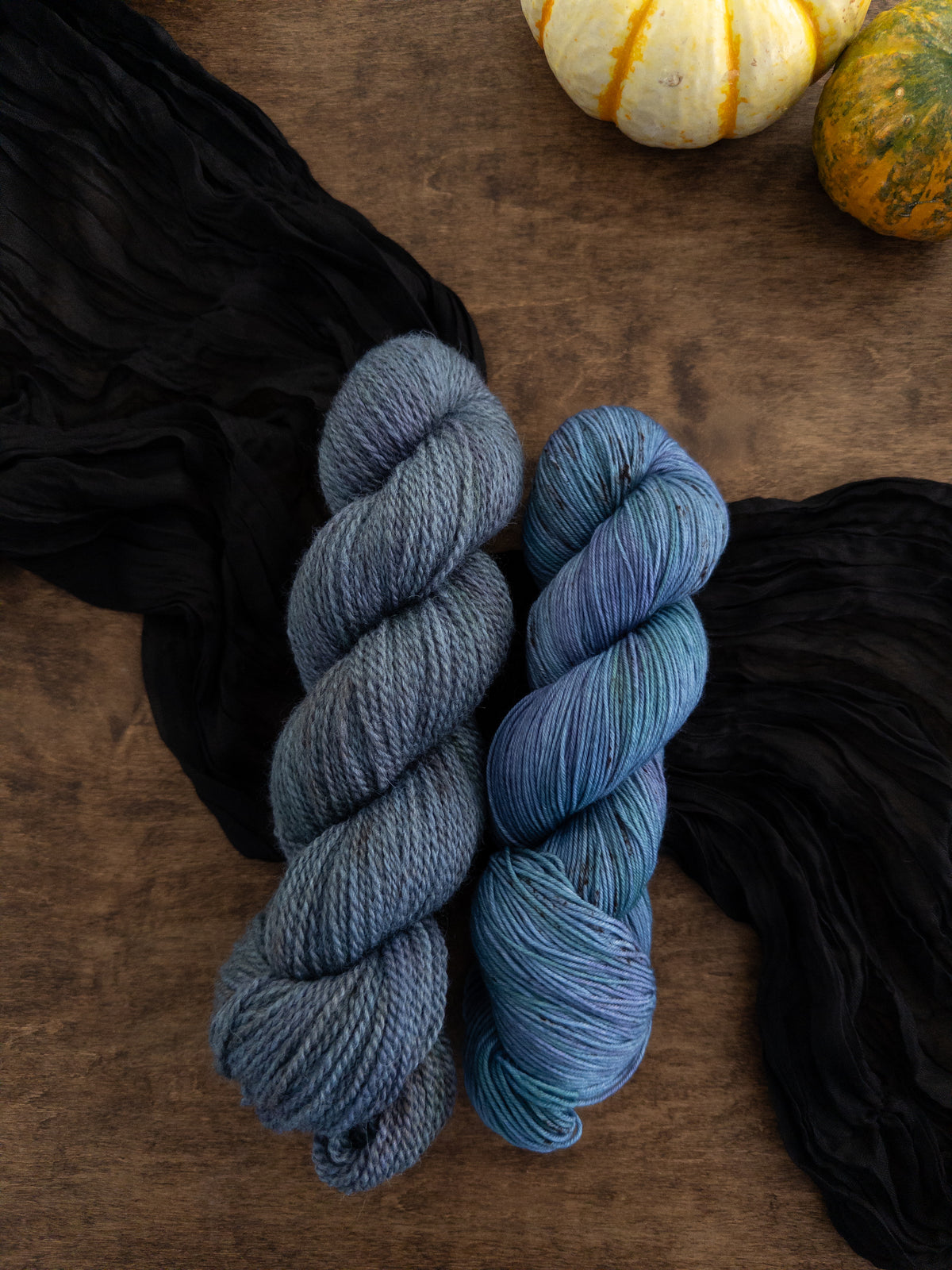 Three skeins of variegated yarn are arranged on a worn wooden table with a strip of black cloth draped diagonally beneath them. The yarn is a cornflower blue with hints of forest green and purple scattered throughout.
