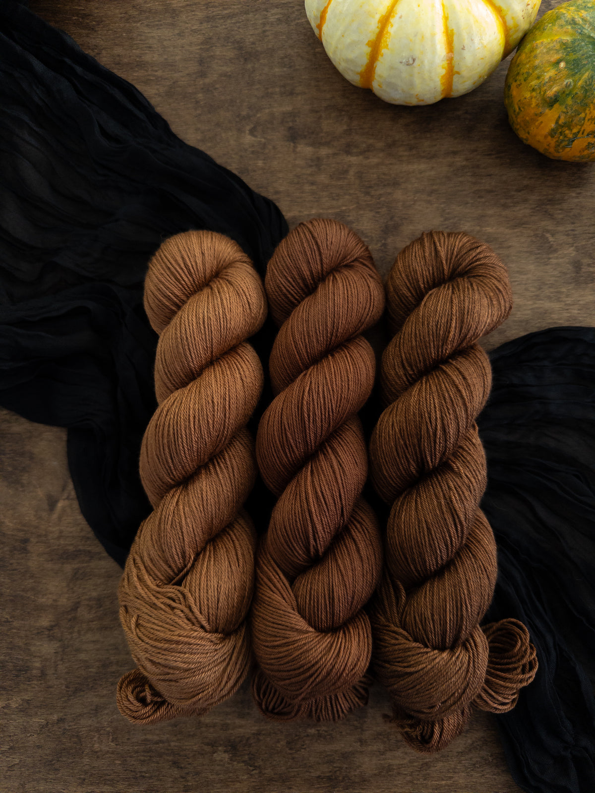 Three skeins of toasty chestnut brown yarn are arranged on a worn wooden table with a strip of black cloth draped diagonally beneath them.