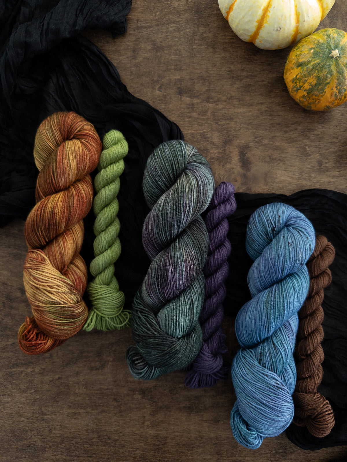 3 sock sets first is a skein that is gold red & brown with a green mini skein Next is a deep forest green with pops of gray & purple with a cool toned purple mini skein The final is a cornflower blue with tones of purple & green with a brown mini skein  