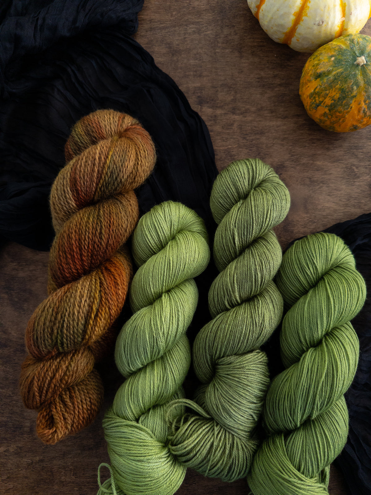 Four skeins of yarn are arranged on a worn wooden table with a black strip of fabric beneath them. The skein to the left is a rustic texture and is variegated with shades of gold, brown, red, and green. The three skeins to its right are a crisp apple green.