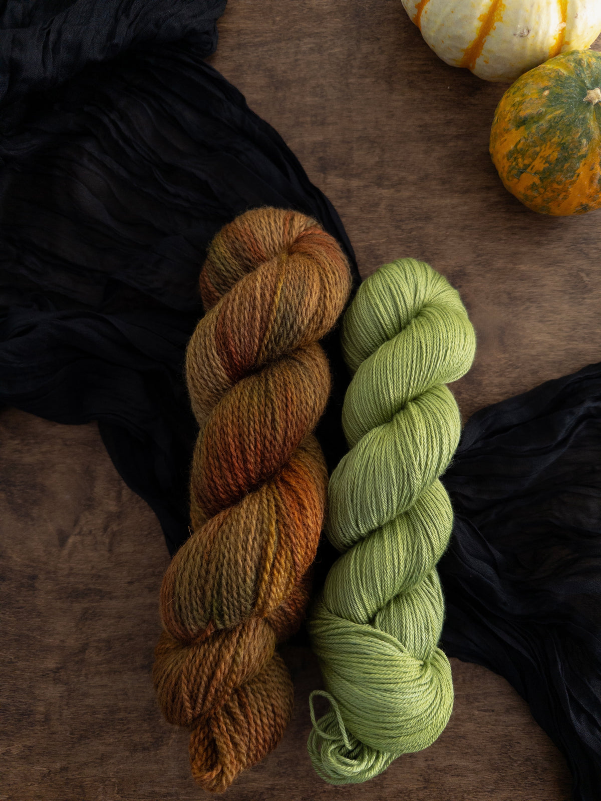 Two skeins of yarn are arranged on a worn wooden table with a black strip of fabric beneath them. The skein to the left is a rustic texture and is variegated with shades of gold, brown, red, and green. The smooth and shiny skein to its right is a crisp apple green.