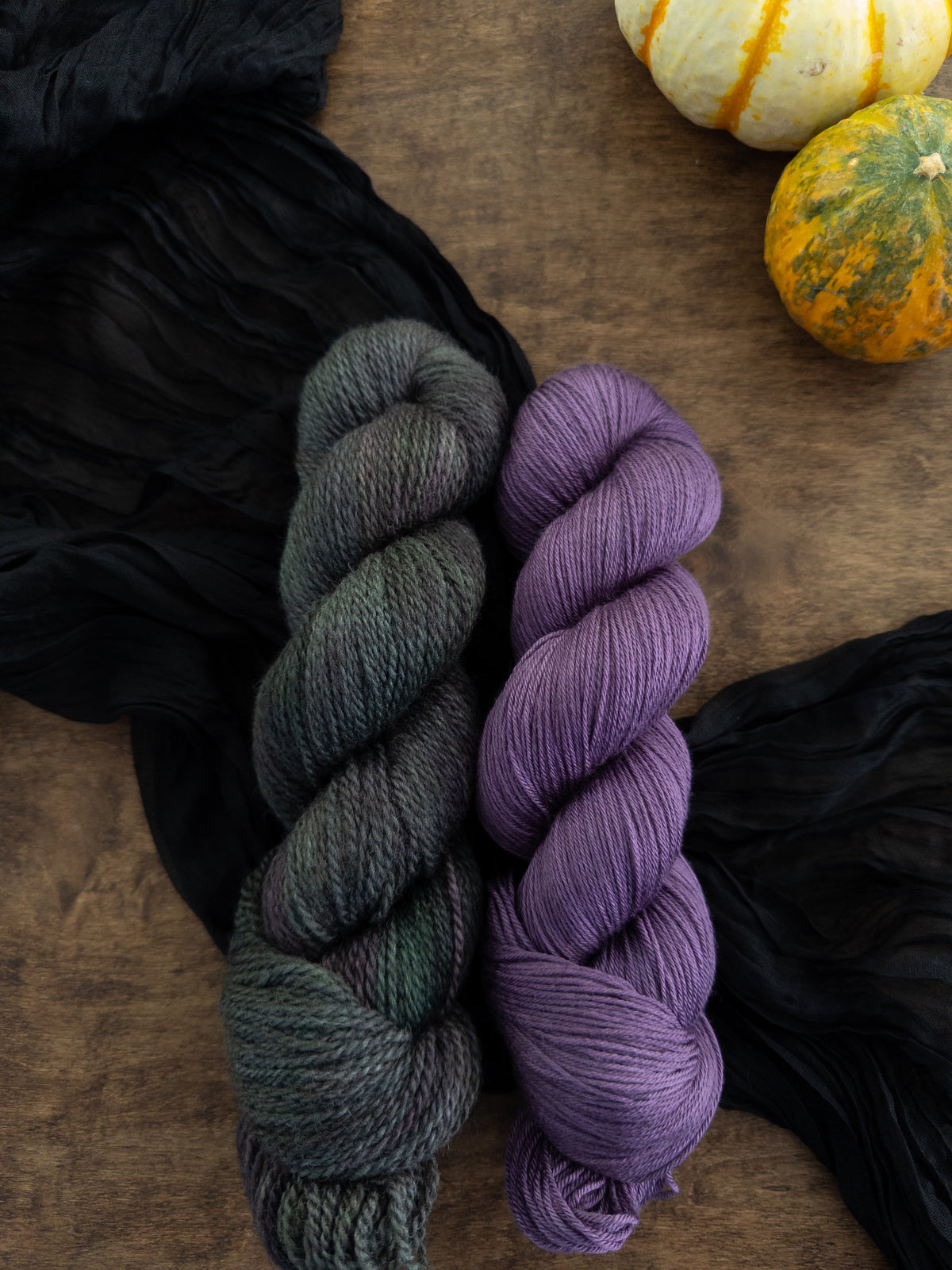 Two skeins of yarn are arranged on a worn wooden table with a black strip of fabric beneath them. The skein to the left is a rustic texture and is variegated with a deep forest green, splashes of gray, lichen, and dark purple. The smooth and shiny skein to its right is a deep, cool toned berry purple.