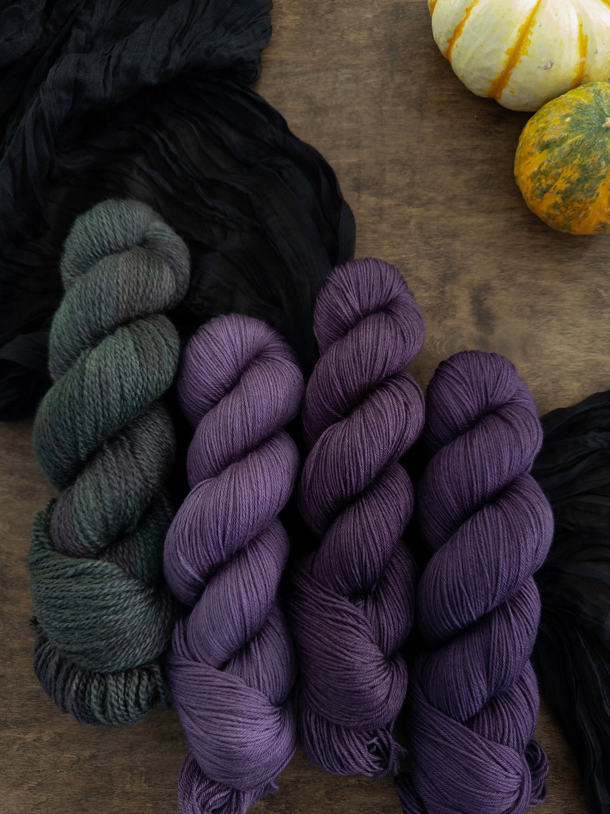 Four skeins of yarn are arranged on a worn wooden table with a black strip of fabric beneath them. The skein to the left is a rustic texture and is variegated with a deep forest green, splashes of gray, lichen, and dark purple. The three skeins to its right are a deep, cool toned berry purple.