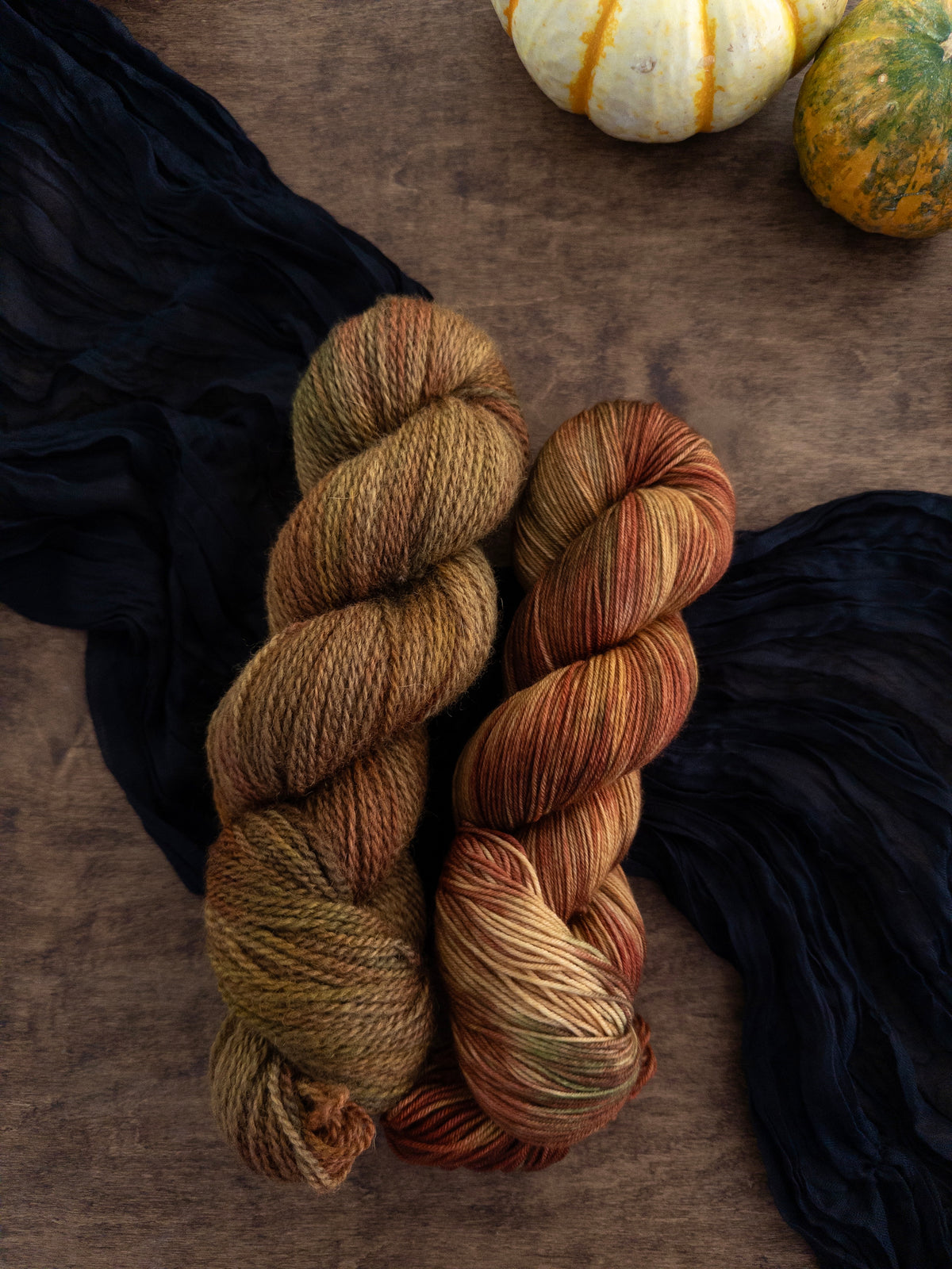 Three skeins of variegated yarn are arranged on a worn wooden table with a strip of black cloth draped diagonally  beneath them. The yarn is a swirl of shades of gold, red, brown, and green like early autumn leaves.