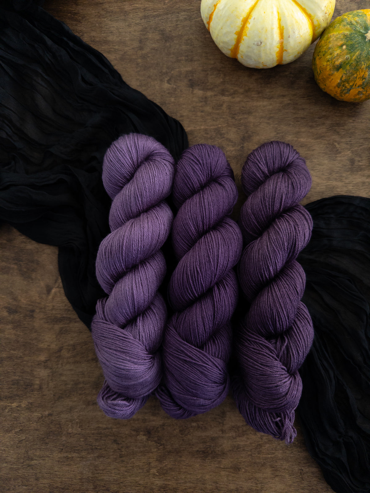 Three skeins of dark cool toned purple yarn are arranged on a worn wooden table with a strip of black cloth draped diagonally  beneath them.