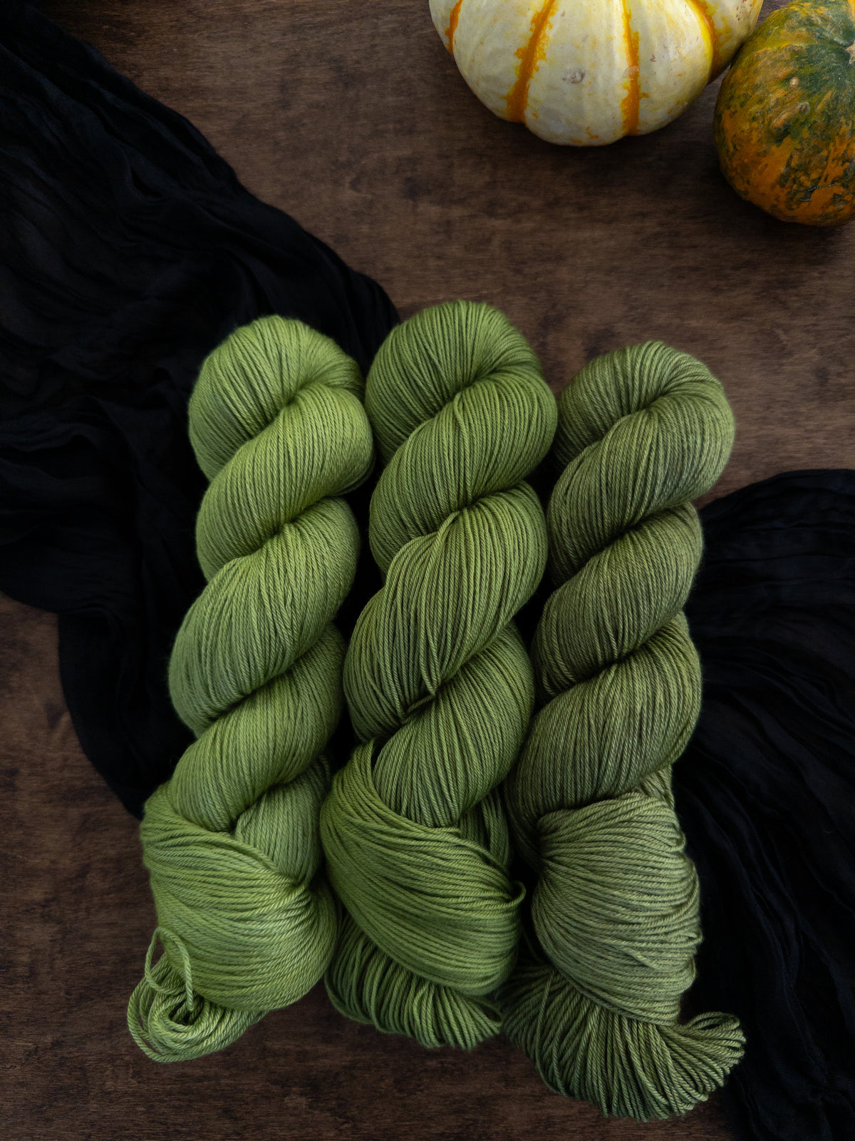 Three skeins of crisp apple green yarn are arranged on a worn wooden table with a strip of black cloth draped diagonally  beneath them.